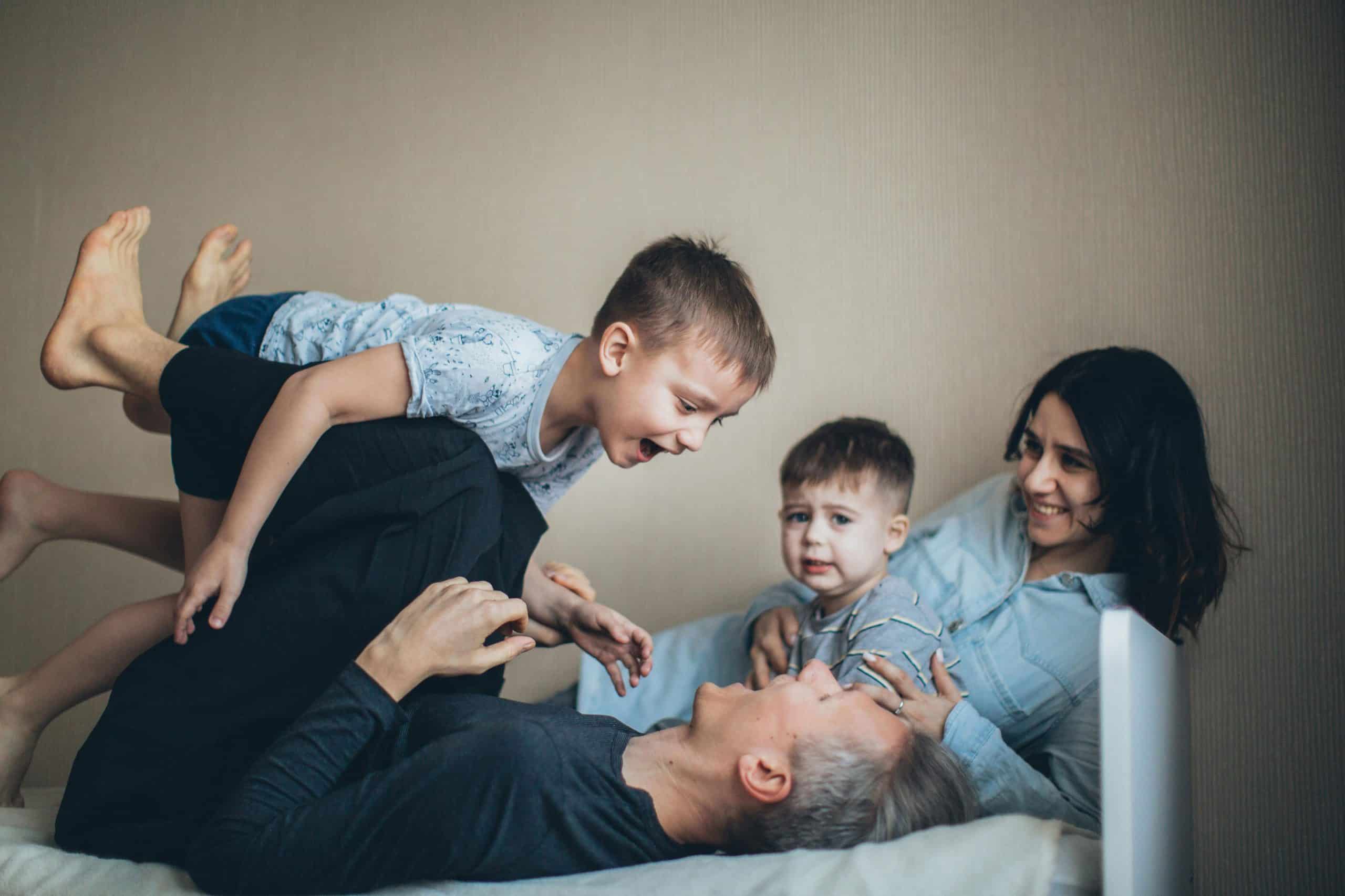 A family laying in bed playing. The dad is lifting a child by his feet. The mom and other child are smiling and looking at them,
