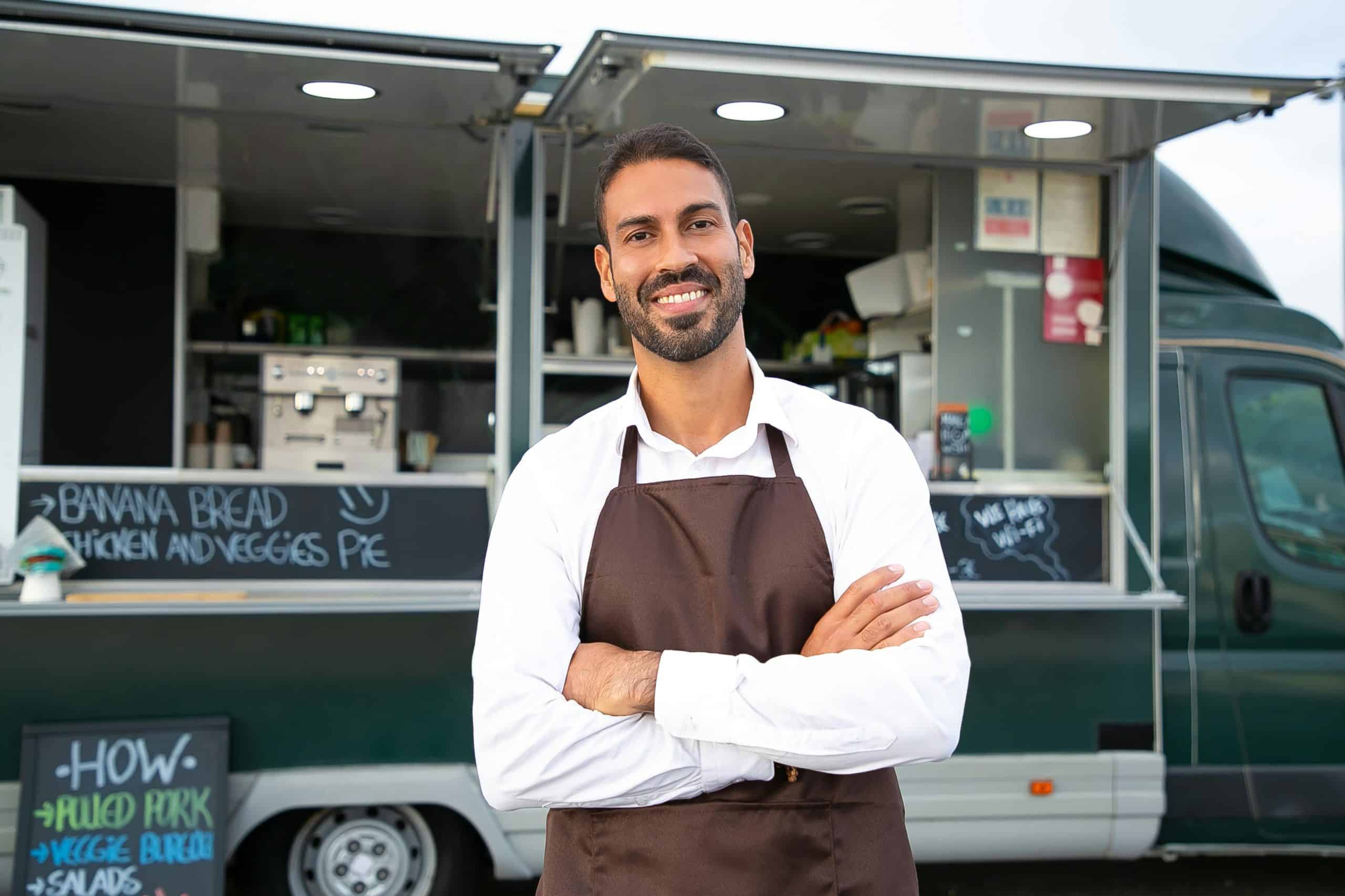 A smiling food truck owner standing in front of his food truck.