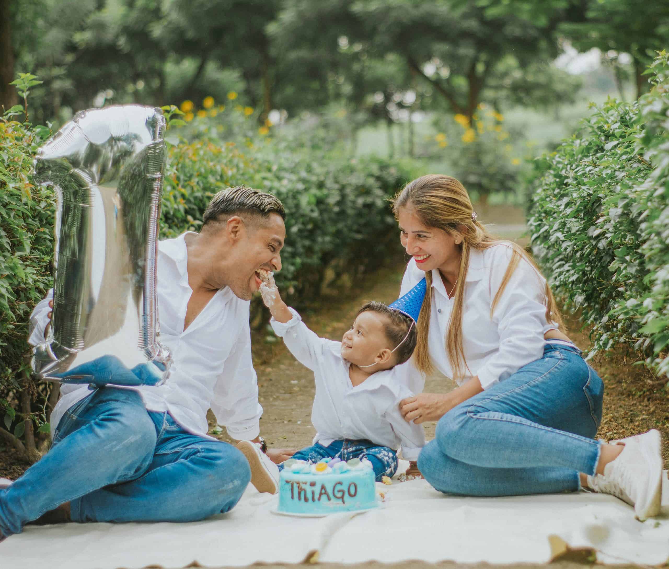 A mom and dad celebrating their one-year old child's birthday on a picnic blanket in a garden. The dad has a mylar balloon shaped as a number 1.