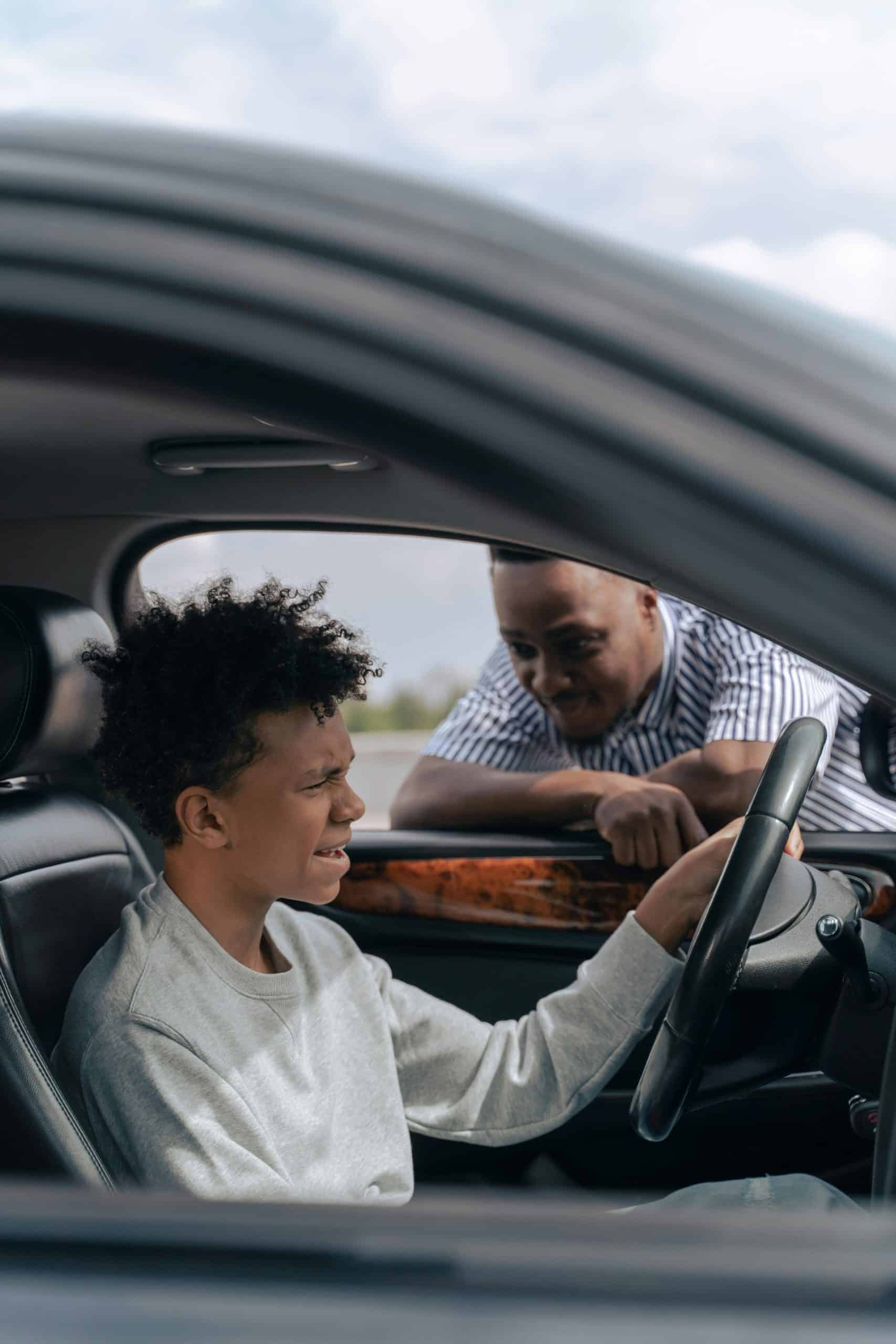 A teenage boy sitting behind the wheel of a car while his father leans in through the driver’s side window.